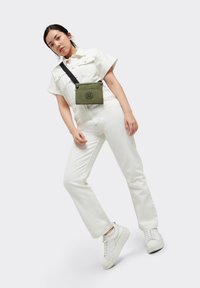 Khaki tvärs över axeln-väska med zip-stängning och logodetalj, bärs över en vit jumpsuit, tillsammans med vita sneakers. Enkelt, funktionellt design.