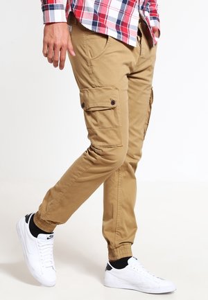 Mand iført beige cargobukser med knaplukning på sidelommer, en rød og blå ternet skjorte, sorte strømper og hvide Nike-sneakers i midtsteget.
