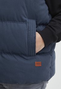 Temno modra podložena brezrokavnik z teksturiranim zaključkom, stranskimi žepi in rjavim usnjenim logotipom. Pod njim je viden črn pulover.