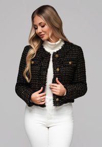 Frau mit langen blonden Haaren, die eine schwarze Tweedjacke mit goldenen Knöpfen über einem weißen Rollkragenpullover und weißen Hosen trägt, nach unten blickt und die Jacke hält.