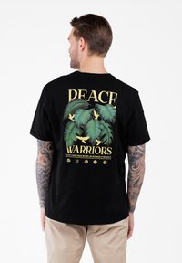 Mężczyzna z tatuażami na ramionach, ubrany w czarny T-shirt z zielonymi liśćmi, żółtymi ptakami i napisem "Wojownicy Pokoju", oraz beżowe spodnie, odwrócony plecami.