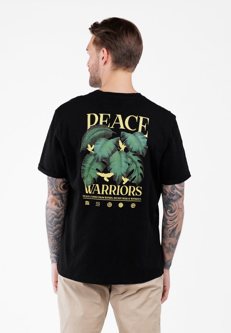 Mężczyzna z tatuażami na ramionach, ubrany w czarny T-shirt z zielonymi liśćmi, żółtymi ptakami i napisem "Wojownicy Pokoju", oraz beżowe spodnie, odwrócony plecami.
