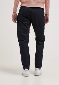 Czarne spodnie cargo o dopasowanym kroju, z gładką powierzchnią materiału, bocznymi kieszeniami i elastycznymi mankietami, noszone z białymi trampkami.