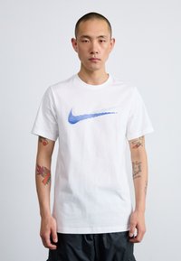 White cotton t-shirt with a blue Nike swoosh logo, short sleeves, round neck, and a relaxed fit, worn over dark shorts.
