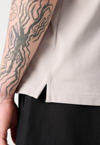 T-shirt en tissu texturé beige clair avec des fentes latérales à l'ourlet, présenté à côté d'un vêtement noir et d'un bras tatoué partiellement visible.