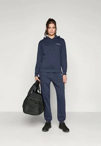 Γυναίκα που στέκεται σε ναυτικό φούτερ και φόρμες, κρατώντας μια μαύρη τσάντα γυμναστηρίου, φορώντας μαύρα αθλητικά παπούτσια σε απλό ανοιχτό φόντο.