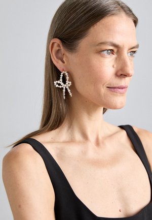Pendientes de perlas blancas en forma de mariposa, con lazos entrelazados y una superficie texturizada, sobre un fondo neutro.