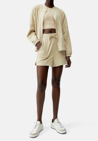 Conjunto atlético beige que incluye una chaqueta bomber con cremallera, un crop top ajustado y shorts con cordón. Tejido suave con un ajuste relajado. Zapatillas blancas.