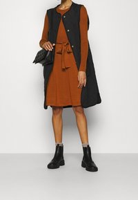 Svart quiltad väst lager på lager över en orange ribbad tröja och matchande klänning med knytskärp, tillsammans med svarta anklar stövlar och en quiltad väska.