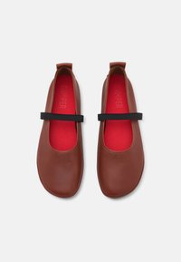 Zapatos de cuero marrón con punta redondeada, interior rojo y tira negra sobre el empeine. Textura suave con un detalle mínimo.