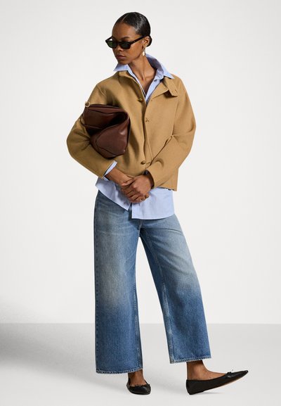 Καφέ σακάκι από μείγμα μαλλιού με κλείσιμο με κουμπιά, φορεμένο πάνω από ένα ανοιχτό μπλε πουκάμισο. Μπλε τζιν παντελόνι με φαρδιά πόδια και μαύρα μπαλαρίνες ολοκληρώνουν την εμφάνιση.