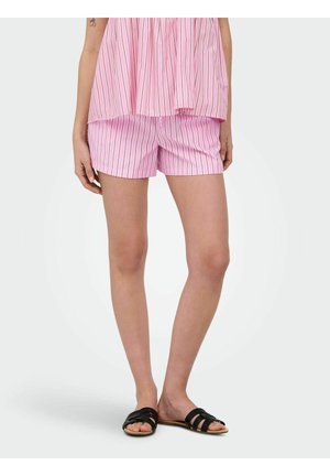 Kobieta w różowo-białych, paskowanych szortach i dopasowanym luźnym topie, w czarnych klapkach, stojąca na jednolitym tle.