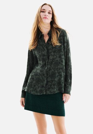 Chemise boutonnée vert foncé avec un motif floral subtil, associée à une jupe mini texturée de couleur vert foncé. Tissu doux avec des manches longues.