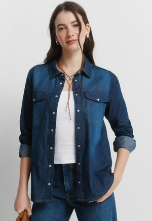 Nuori nainen pukeutuneena tummaan denimpaitaan valkoisen topin päällä, samaa farkkumateriaalia olevat farkut, pitkät hiukset ja kerrokselliset kaulakorut, poseeraa käsi taskussa.