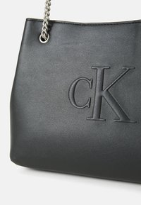 Czarna skórzana torebka z wytłoczonym logo "CK", teksturowa powierzchnia i srebrny łańcuszek. Posiada otwarte dno i zarysowaną formę.