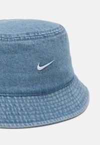 Bucket hat made of light blue denim, featuring a white embroidered Nike logo. Textured fabric with a wide, structured brim.