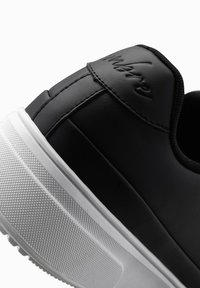 Baskets noires avec une tige en cuir lisse, des détails en relief sur le talon, et une semelle blanche texturée pour une bonne adhérence. Silhouette incurvée.