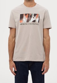 Muž v béžovém tričku Armani Exchange s barevným abstraktním logem AX a černými džíny na bílém pozadí.