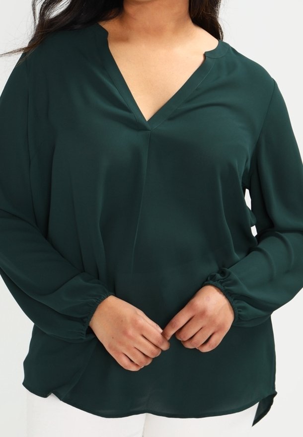Femme portant un chemisier vert foncé à manches longues avec un col en V et des poignets élastiques, les mains jointes devant, associée à un pantalon blanc.