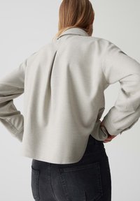 Helle graue, taillierte Jacke mit umgeschlagenem Kragen, langen Ärmeln und einer Rückenfalte, kombiniert mit schwarzen Denim-Jeans. Glatte Textur, minimalistisches Design.