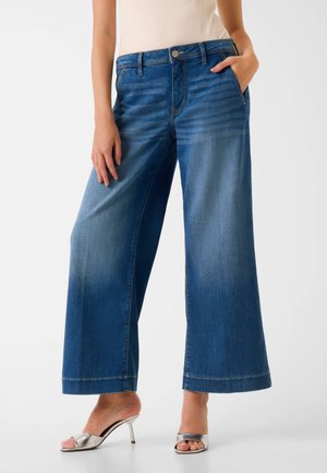 Femme portant un jean bleu à jambes larges, taille haute, bouton sur le devant, et des sandales argentées à talons hauts et orteils ouverts, debout avec une main dans la poche.