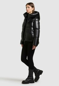 Giacca a vento nera con finitura lucida, cappuccio oversize, due tasche frontali e chiusura con zip. Indossata con leggings neri e stivali militari.