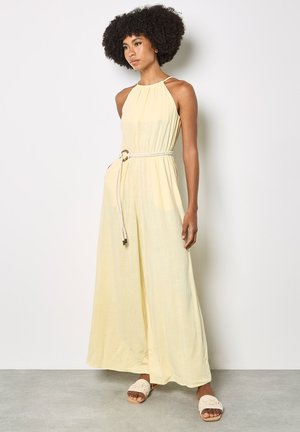 Kobieta o kręconych włosach, ubrana w bezrękawny, jasnożółty kombinezon na szyję z szerokimi nogawkami i białym paskiem z liny, w połączeniu z białymi plecionymi sandałami.