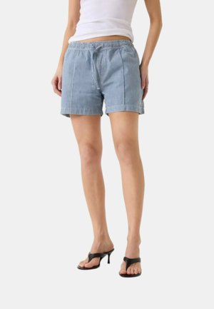 Donna che indossa pantaloncini a righe azzurri con coulisse, una canotta bianca senza maniche e sandali neri con tacco a infradito, in piedi su sfondo bianco.