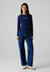 Maglietta a maniche lunghe navy con la scritta "RED BULL RACING" in bianco, abbinata a jeans larghi in denim blu e scarpe bianche con suole marroni.