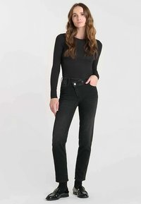 Jean noir taille haute avec poche avant, texture lisse et fermeture à boutons contrastants. Modèle portant un haut noir à manches longues ajusté.