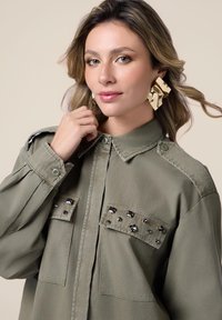 Camicia verde oliva con colletto e bottoni, presenta due tasche sul petto con borchie nere e spalline decorative. Tessuto morbido.