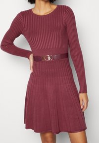 Robe en maille côtelée bordeaux avec un corsage ajusté et une jupe évasée. Comprend des manches longues et une ceinture brillante à la taille avec une boucle dorée.