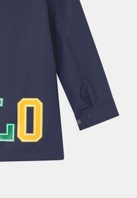 Námořnická nepromokavá bunda s manžetami na knoflíky, na přední straně s odvážným zeleným a žlutým textem v kontrastních barvách.