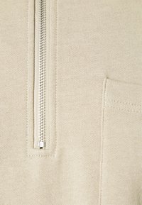 Gros plan sur un tissu beige avec une fermeture éclair argentée et une poche rectangulaire cousue sur le côté droit.