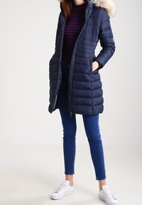 Donkerblauwe pufferjas met een bontgevoerde capuchon, gewatteerd ontwerp, rits aan de voorkant en zijzakken, gecombineerd met blauwe spijkerbroeken en witte sneakers.