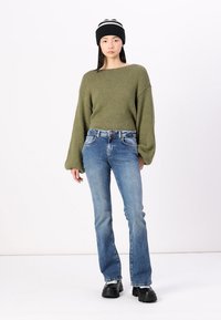 Olivgrön texturerad tröja med ballongärmar, blå slim fit-jeans och svart mössa med vita ränder, kombinerat med svarta chunky boots.