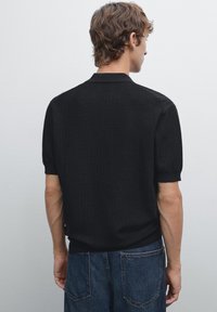 Polo shirt nero a costine con maniche corte, dotato di un colletto classico e una vestibilità aderente all'orlo, abbinato a jeans in denim scuro.