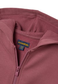 Sudadera con cremallera en un suave color malva, fabricada con un tejido suave y forro texturizado. Presenta una etiqueta tejida con "essentials" en azul y amarillo.