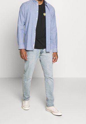 Světle modrá košile se zapínáním na knoflíky přes černé tričko se žlutým grafikou, v kombinaci se světle modrými džíny a bílými teniskami.