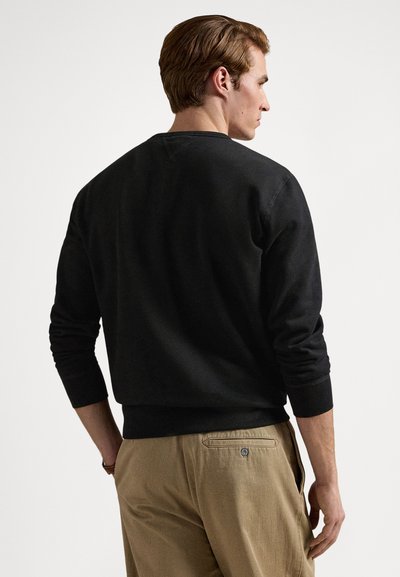 Μαύρο φούτερ με στρογγυλή λαιμόκοψη και πλεκτές μανσέτες, συνδυασμένο με ανοιχτό μπεζ παντελόνι με πλαϊνές τσέπες και τσέπη με κουμπί στο πίσω μέρος.