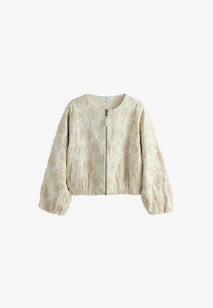 Mujer con chaqueta bomber bordada con flores en color crema y shorts con borde de encaje, de pie cerca del océano con cielo nublado de fondo.