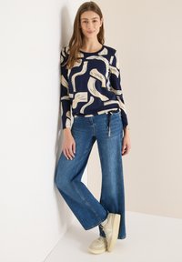 Nainen pukeutuneena laivastonsiniseen pitkähihainen toppiin, jossa on beigejä abstrakteja kuvioita, sinisiin leveälahkeisiin farkkuihin ja kermaisiin tennareihin, nojaa valkoiseen seinään.