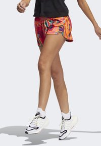 Mujer con pantalones cortos de correr coloridos y zapatillas Adidas blancas en medio de un paso contra un fondo simple.