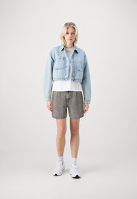 Veste en denim bleu clair courte, T-shirt blanc et shorts gris au toucher lisse. Des chaussures de sport blanches et des chaussettes de cheville complètent la tenue.