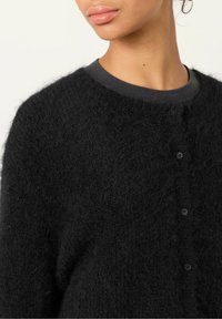 Svart, fluffig cardigan med rundad halsringning och mjuk textur, med en knappstängning fram och ett enhetligt mörkgrått innerfoder.