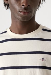 Hvid bomulds t-shirt med marineblå vandrette striber, ribbet krave og lille broderet logo på brystet. Nærbillede af stofstrukturen.