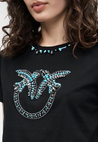 Kvinne iført svart skjorte med to perlede fugler i turkis og sølvstein som danner et sirkelmønster foran.