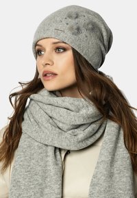 Szary dzianinowy beanie z dekoracyjnymi futrzanymi pomponami i cyrkoniami; miękki szary szalik zarzucony na szyję, oba wykonane z teksturowanej tkaniny.