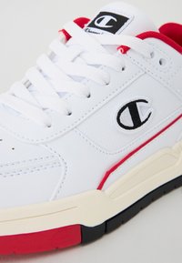 Valged sneakers tekstureeritud nahast pealsel, punaste aktsentidega ja musta logoga. Omadused hõlmavad valgeid kingapaelu ja kummist talla kreemjas ja mustas värvitoonis.