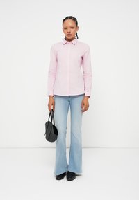 Camisa de botão riscada em rosa e branco com colarinho clássico, combinada com jeans flare azul claro. Segurando uma pequena mala preta.